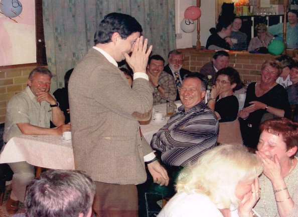 Mr Bean imitator fluistert in oor, terwijl feestende mensen aan tafel lachen in een drukke zaal.