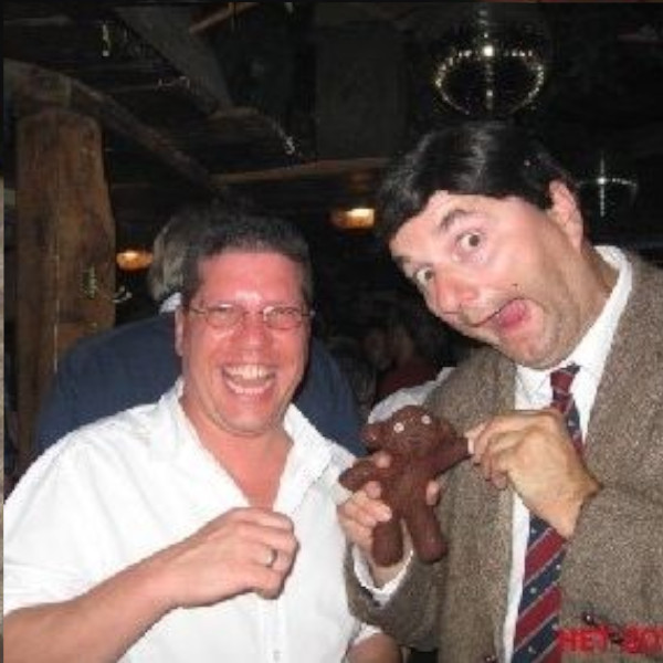 Twee mannen lachen in een feestelijke setting, terwijl één de teddybeer van Mr Bean lookalike vasthoudt.